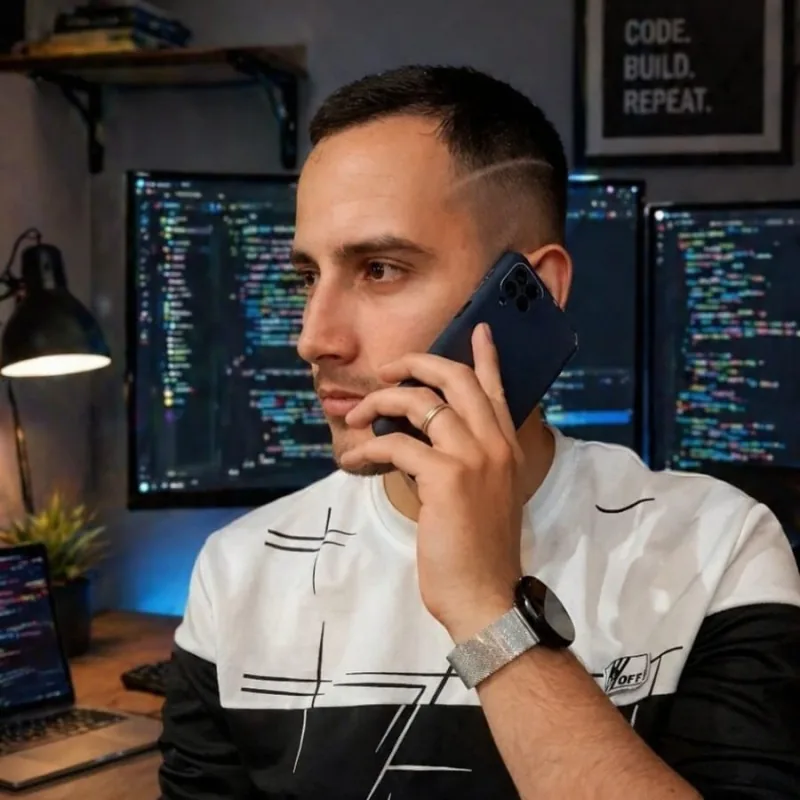 Gabriel, de camiseta preta e branca, falando ao telefone em frente a monitores com código.